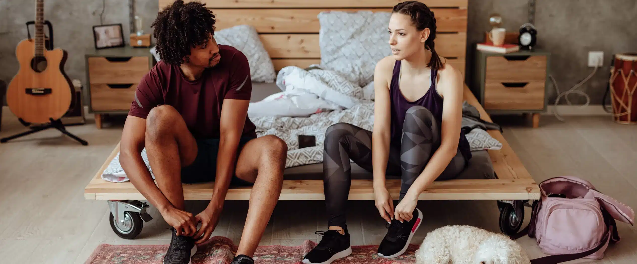 a couple sits on the edge of a bed, tying their gym shoe laces before a workout