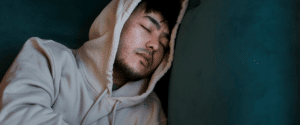 a man wears a hooded sweatshirt and sleeps sitting up with his head against the wall