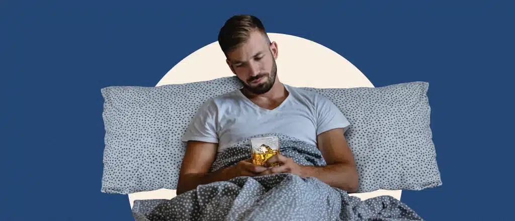 a tired-looking man sits upright in bed holding a glass of whiskey
