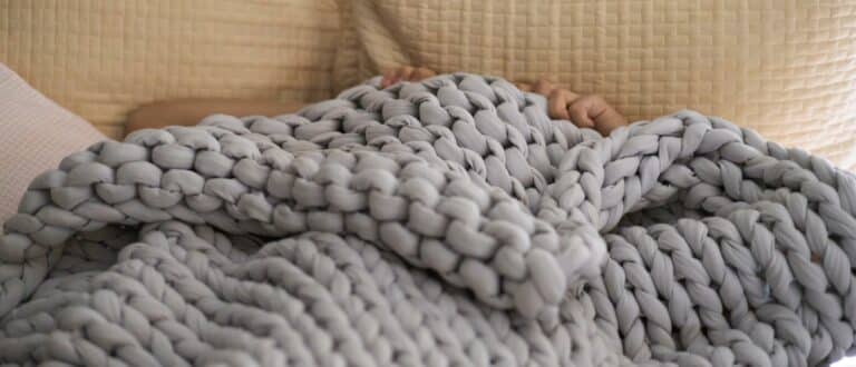 a man pulls a knit weighted blanket up over his face while lying in bed