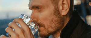 a close up of a man drinking whiskey out of a crystal glass and looking into the distance