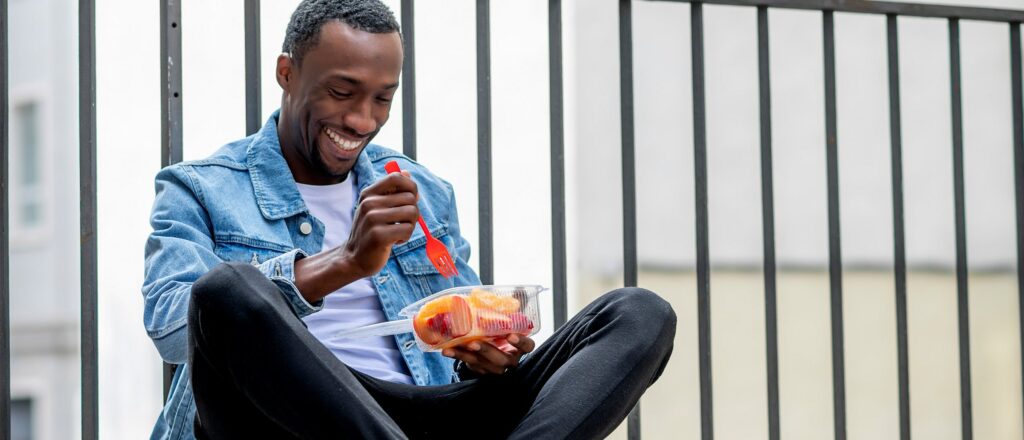 a man sits outside with his legs crossed, smiling while eating a fruit bowl