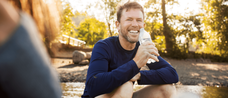 a man sits outside in nature smiling while sipping a water bottle