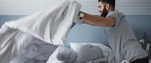 A man stands next to bed and fluffs sheets