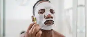 A man wearing a sheet mask looks into a mirror