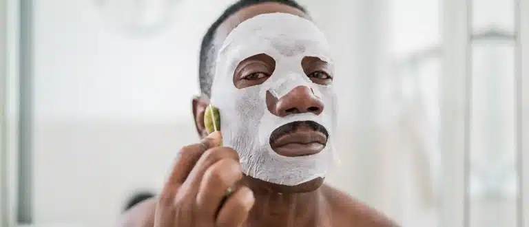 A man wearing a sheet mask looks into a mirror