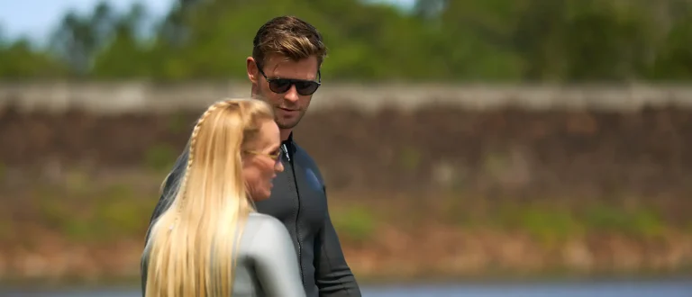 Chris Hemsworth looking at Tanya Streeter while standing on a dock in wetsuits.