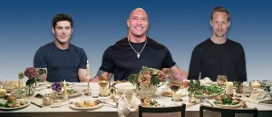 Zach Efron, Dwayne Johnson, and Aleksander Skarsgard sitting at a table covered in loads of food.