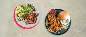 One plate with salmon and a salad next to a plate with a burger and fries