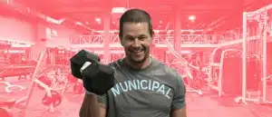 Mark Wahlberg holding a heavy dumbbell in front of a gym in a Municipal t-shirt.