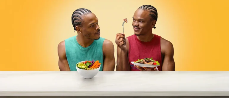 two twins from you are what you eat looking at each other with plant-based food on one's plate, and meat on the other.