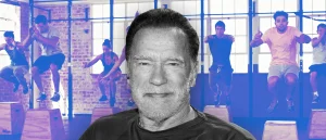 Arnold Schwarzenegger smiles in front of a gym full of people doing box jumps.